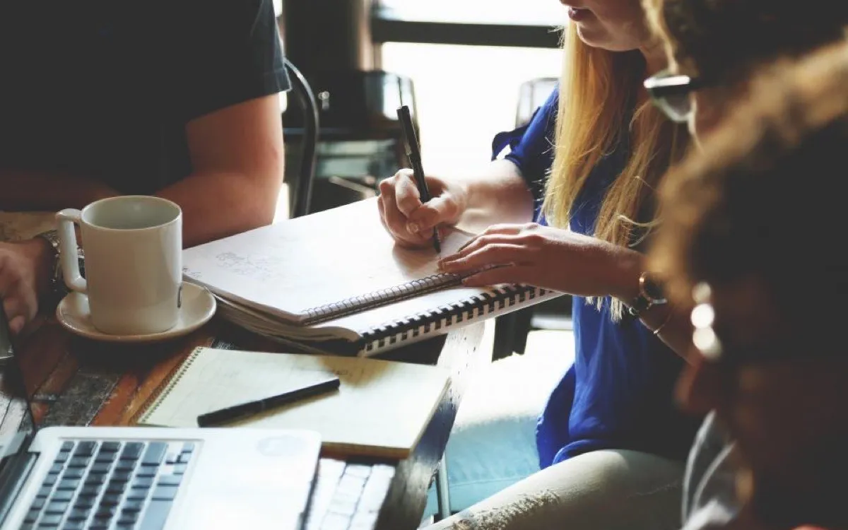 Someone writing in a notebook at a desk beside an open laptop and a coffee