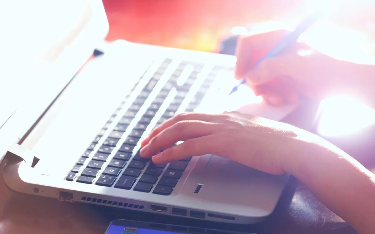 Hands typing on a laptop keyboard, backlit by a window