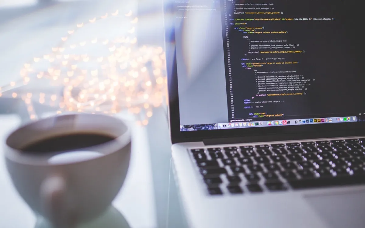 A laptop showing code beside a cup of coffee, warm string lights behind