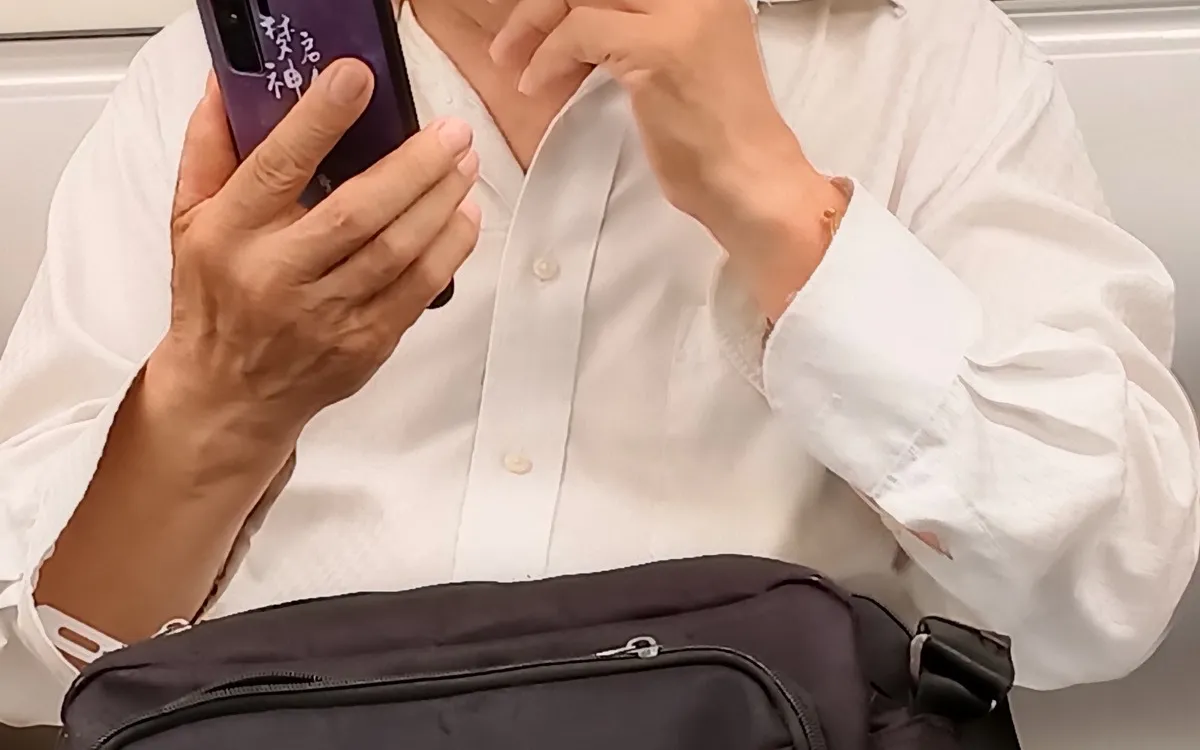 A person in a white shirt holding a phone in both hands, shoulder bag across the chest